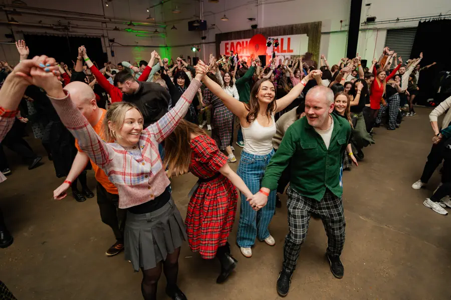 A packed public ceilidh, dancers in tartan with arms raised